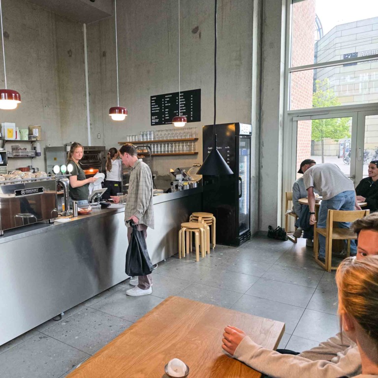 G&aelig;ster ved disken og bordene hos F&aelig;tter p&aring; Godsbanen i Aarhus C, med betonv&aelig;gge og store vinduespartier.