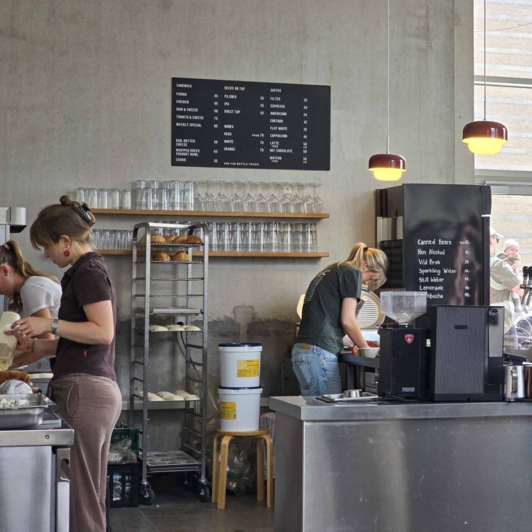 Medarbejdere ved disken hos F&aelig;tter p&aring; Godsbanen, med menutavle og reol med hjemmebagte boller i baggrunden.