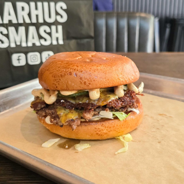 Smash burger med to b&oslash;ffer, smeltet ost og salat foran Aarhus Smash-logoet.