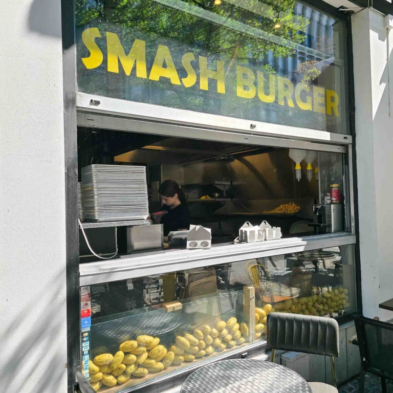 Vindue med "Smash Burger"-logo hos Aarhus Smash, medarbejder ved arbejdet og friske kartofler i vinduet.
